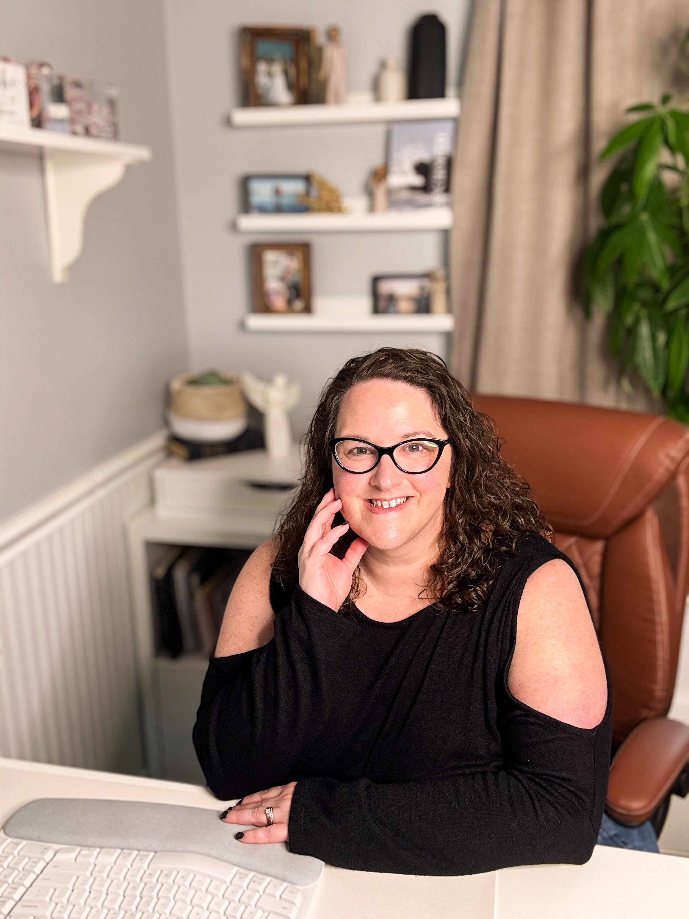 Woman sitting at a desk in an office setting with shelves and decor in the background.